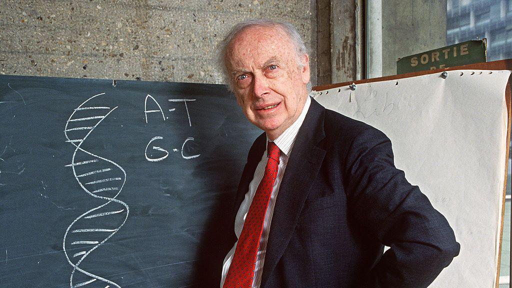 James Watson, de pie junto a una pizarra. Viste una chaqueta oscura, una camisa blanca y una corbata roja.