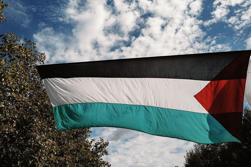 علم فلسطين بالألوان الأخضر والأبيض والأسود والمثلث الأحمر، يرفرف وفوقه سماء زرقاء تظهر فيها سحب بيضاء بكثافة، وعلى جانبي الصورة أشجار خضراء.