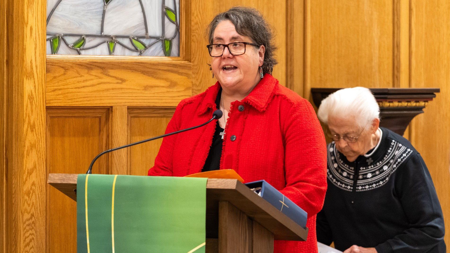 La hermana Susan, vestida con una chaqueta roja y gafas, habla desde un atril cubierto con una tela verde y con una carpeta azul delante. Detrás de ella, una persona mayor mira fijamente algún tipo de lectura.