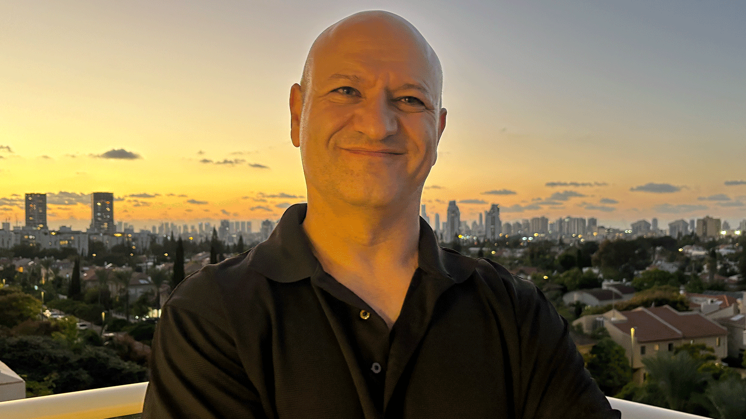 Michael Milshtein em uma sacada, sorri para a c&acirc;mara. Ao fundo, o entardecer e os arranha-c&eacute;us de uma cidade israelense