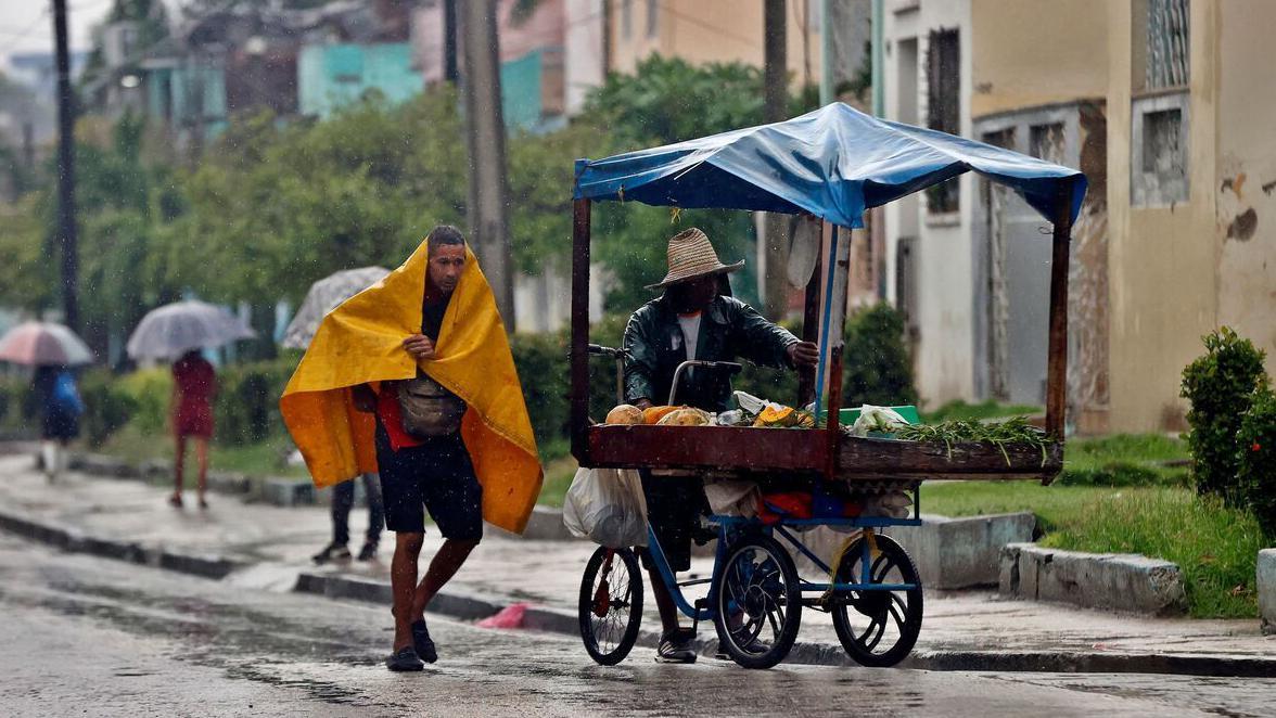 Dvojica muškaraca se sklanjaju od poplave, jedan je pokriven žutom ceradom, dok drugi vozi kolica sa hranom
