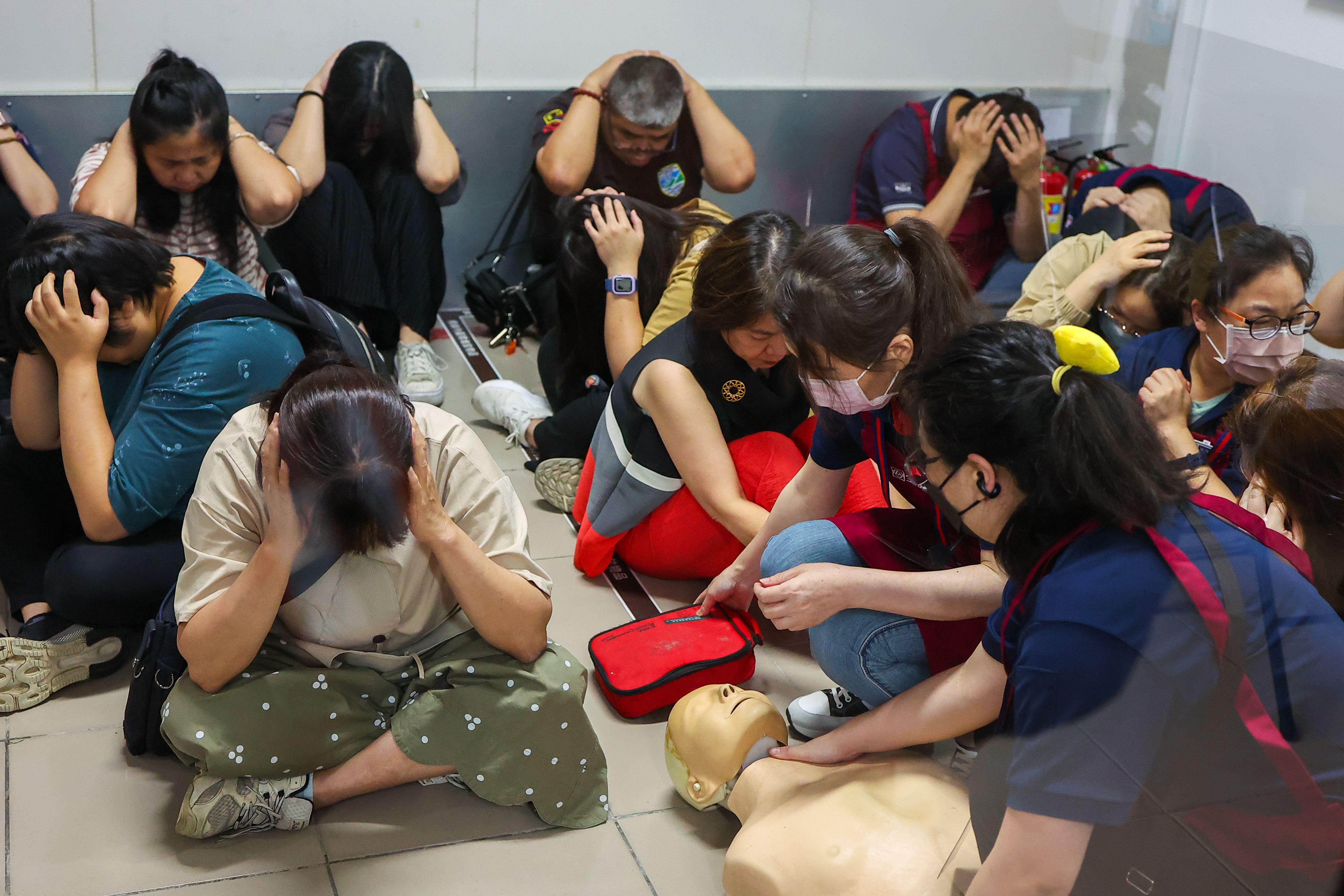 Pessoas sentadas no chão com as mãos na cabeça. Há um boneco no chão e duas pessoas demonstram como fazer atendimento