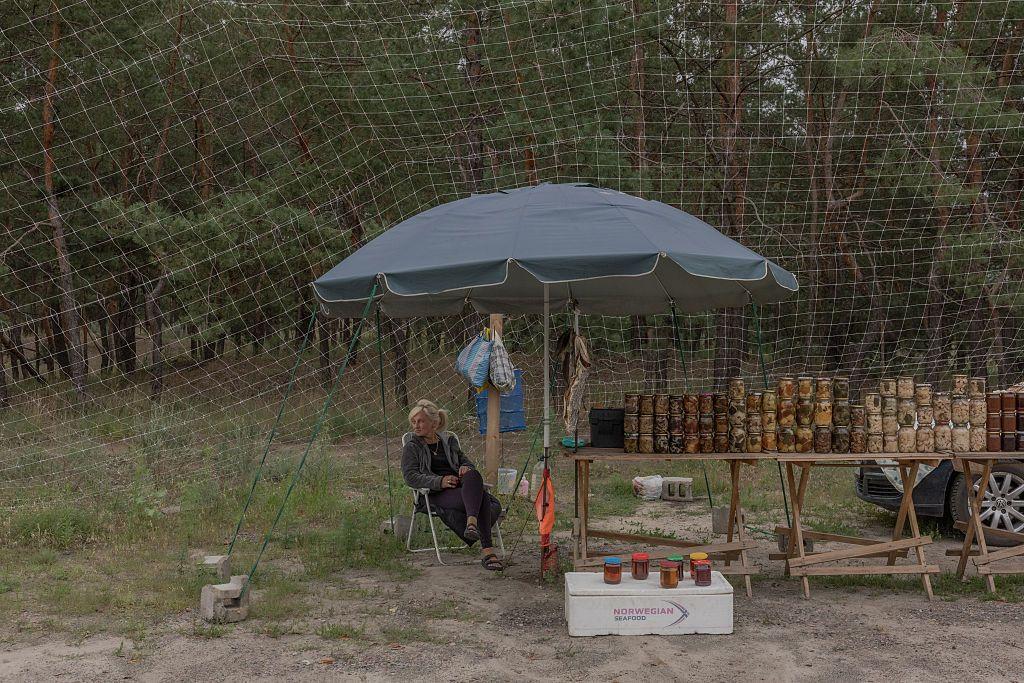 На узбіччі біля соснового лісу облаштовано невеликий придорожній прилавок із домашніми консерваціями. Під великим навісом на столах виставлені десятки банок із закрутками, поруч сидить продавчиня. Над торговим місцем натягнута велика протидронова сітка, яка вкриває всю ділянку.