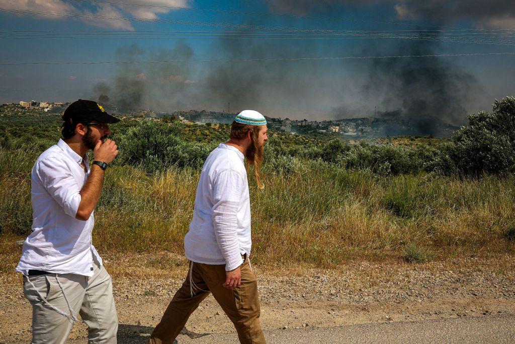 مستوطنان إسرائيليان ينظران إلى الدخان المتصاعد فوق بلدة دوما بالقرب من نابلس في الضفة الغربية المحتلة. يظهر في الصورة التي التقطت نهاراً أعشاب وبعض الأشجار والمباني البعيدة.