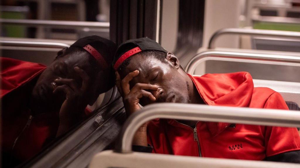 Homem negro dorme sentado em um ônibus. Ele usa em um boné e uma jaqueta vermelha.