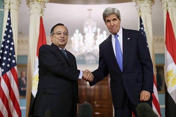 كيري يرتدي بدلة رسمية كحلية اللون وقميصاً أبيض وربطة عنق زرقاء يقف إلى يمين الصورة مصافحاً فهمي الذي يرتدي بدلة رسمية سوداء وقميصاً أبيض اللون وربطة عنق بلون فيروزي. وخلفهما أعلام مصر والولايات المتحدة.