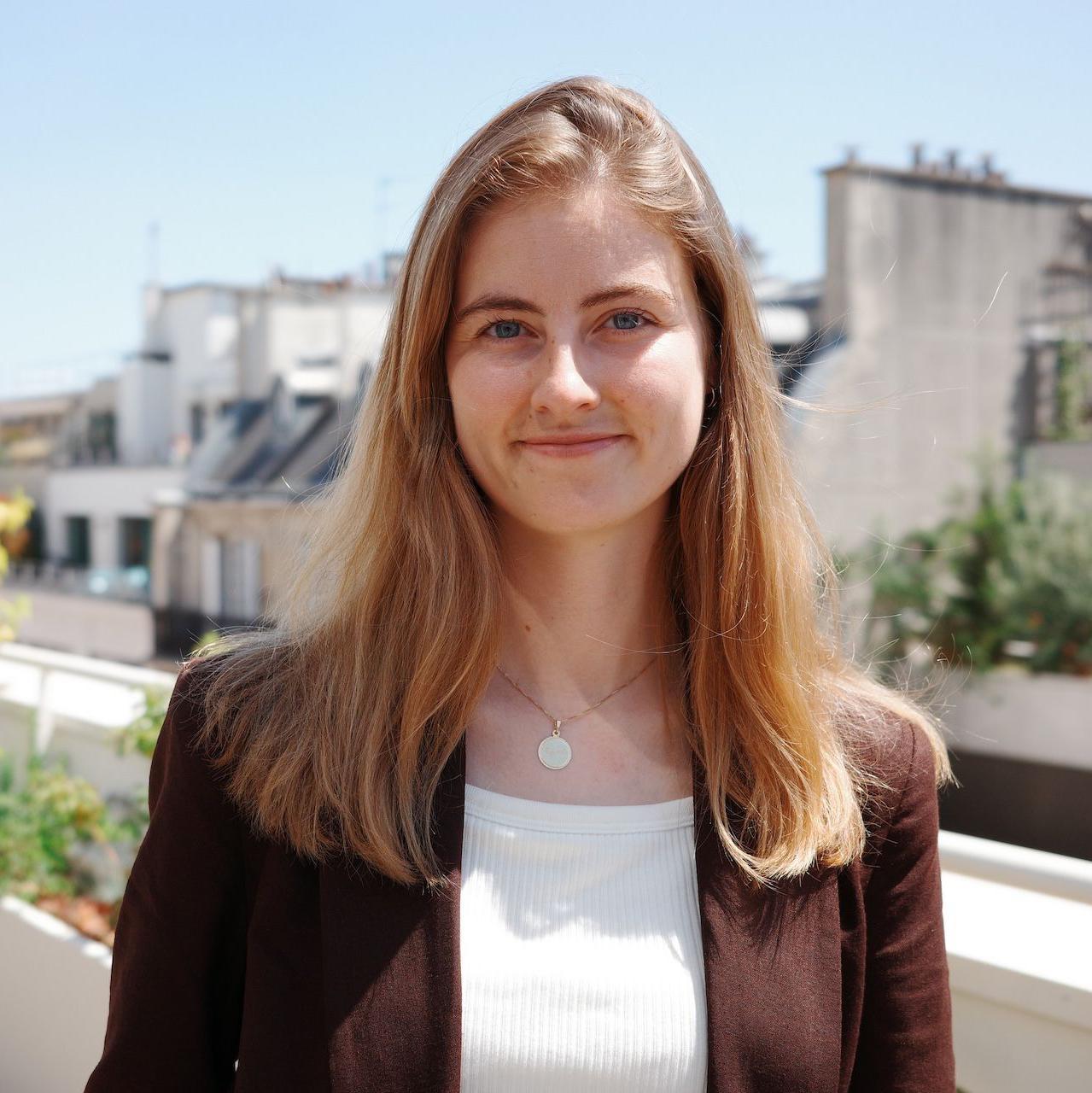 Emma Fourreau est&aacute; em uma varanda ensolarada com plantas ao fundo. Ela veste uma jaqueta marrom aberta e usa uma blusa branca canelada por baixo. Emma Fourreau est&aacute; sorrindo na foto.