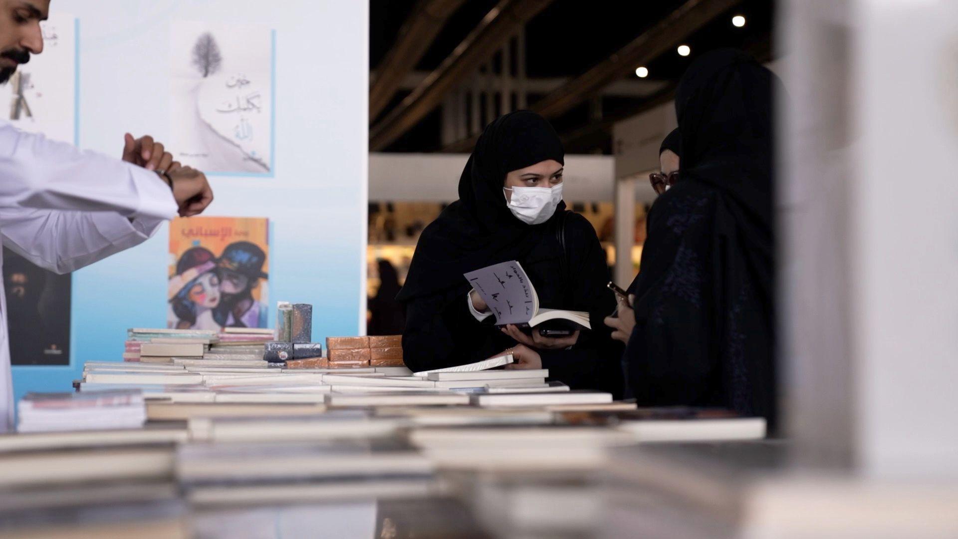 بين الانفتاح والرقابة: معرض الرياض للكتاب يكشف ملامح التحوّل الثقافي السعودي 3 سيدة في المعرض تقف، مرتديةً عباءة سوداء وغطاء شعر أسود، وتغطي وجهها كمامة طبية، بينما تتصفح كتاباً معروضاً، وتتحدث إلى سيدتين تقفان بجوارها، وأمامهم جميعاً طاولة وُضعت عليها كتب كثيرة.