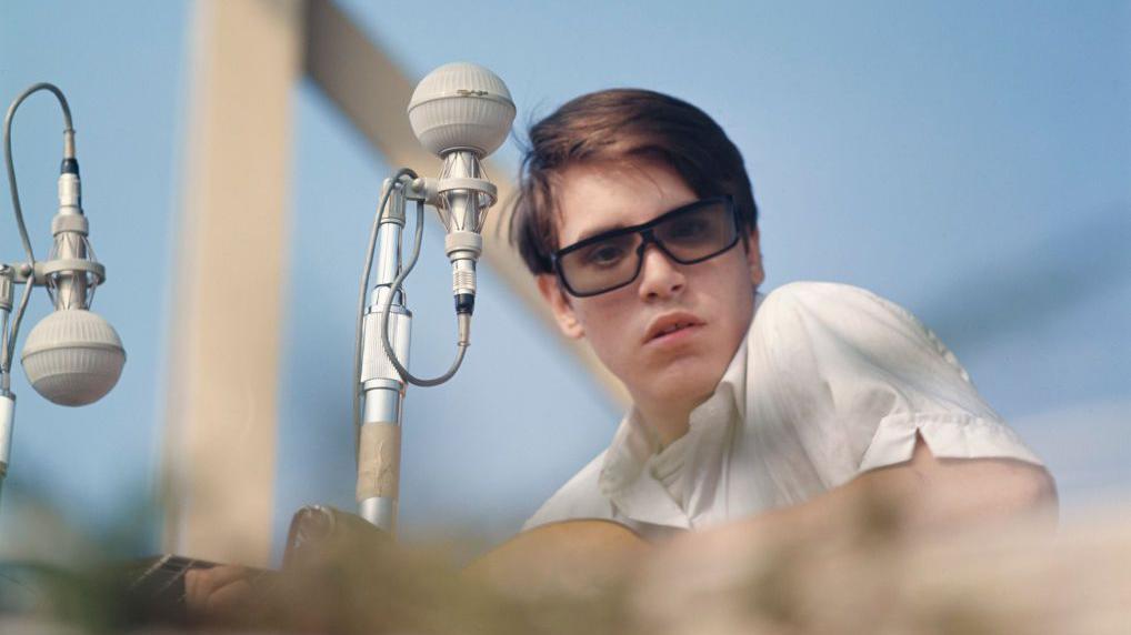 Un joven José Feliciano se presenta en el Newport Folk Festival, en Rhode Island, en 1964. Lleva una camisa de botones blanca, su guitarra en mano y unas gafas oscuras en la cara. Está sentado frente a un micrófono de la época. 