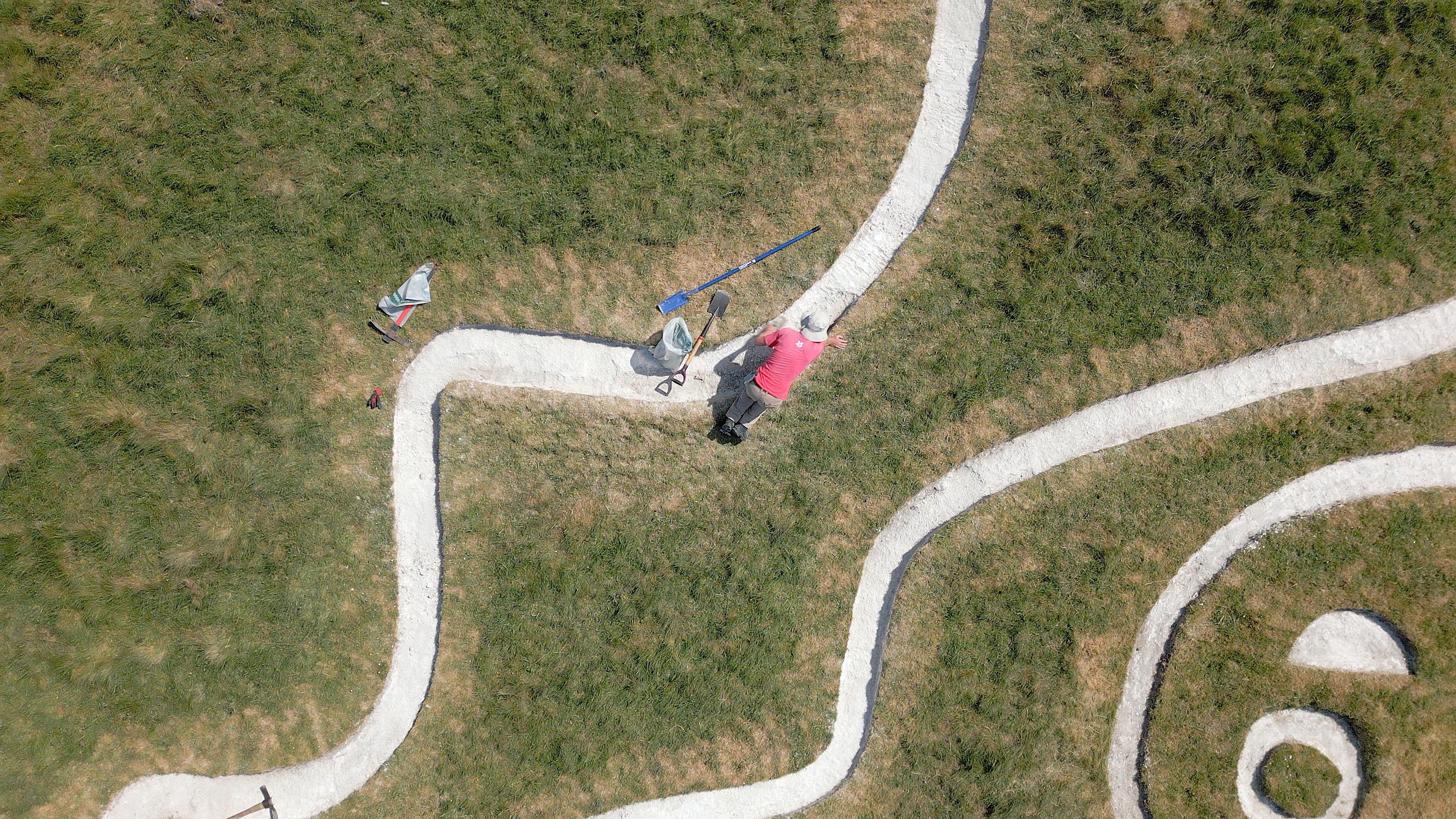 Un hombre con una camiseta roja y un sombrero se ve desde arriba. Está a cuatro patas sobre una línea de tiza blanca, con una pala y una bolsa de plástico a su lado. 