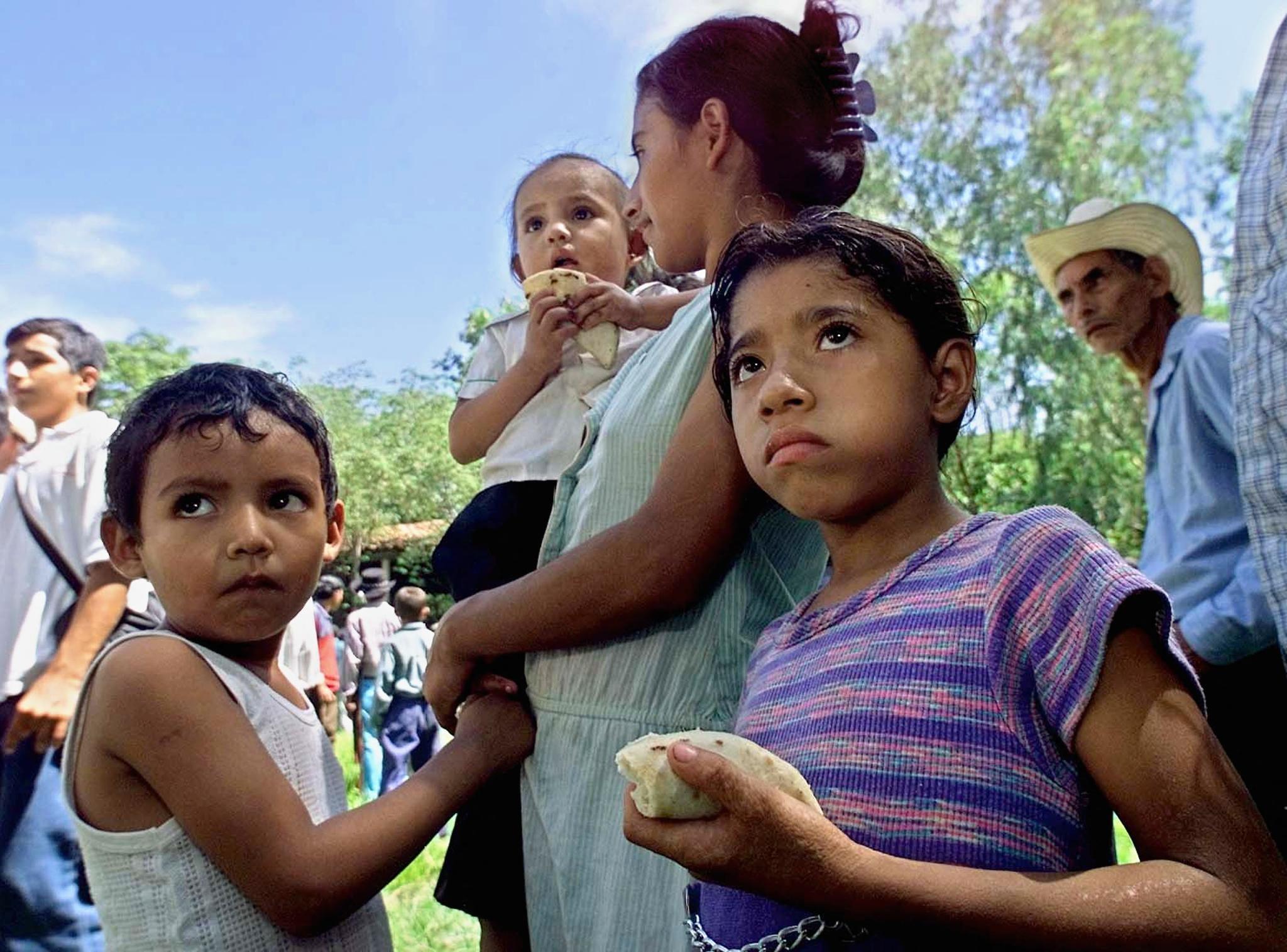 Una mujer carga en brazos a una niña, y hace fila junto a otros dos hijos para recibir recibir raciones de alimentos.