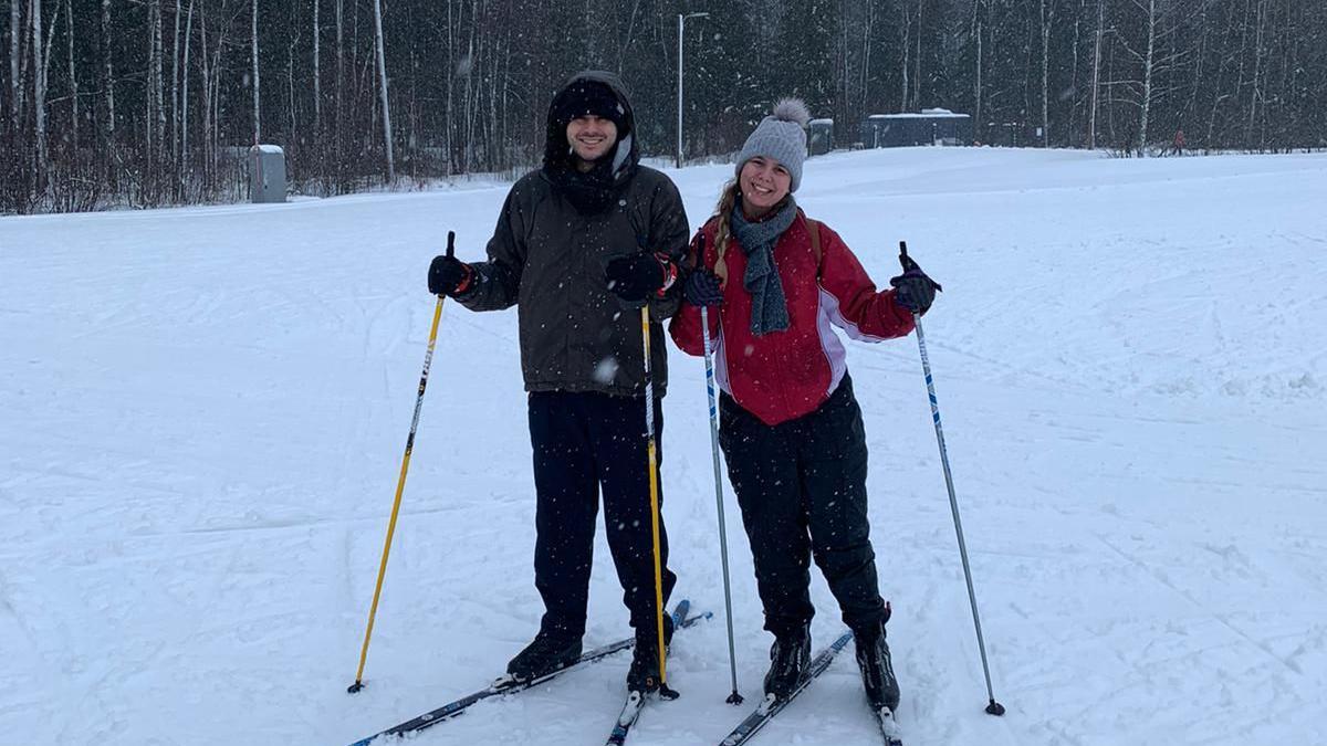 Um casal composto por um homem e uma mulher pratica esqui em meio &agrave; neve em uma &aacute;rea arborizada, vestindo roupas de inverno e segurando bast&otilde;es, em um cen&aacute;rio t&iacute;pico de pa&iacute;ses n&oacute;rdicos.