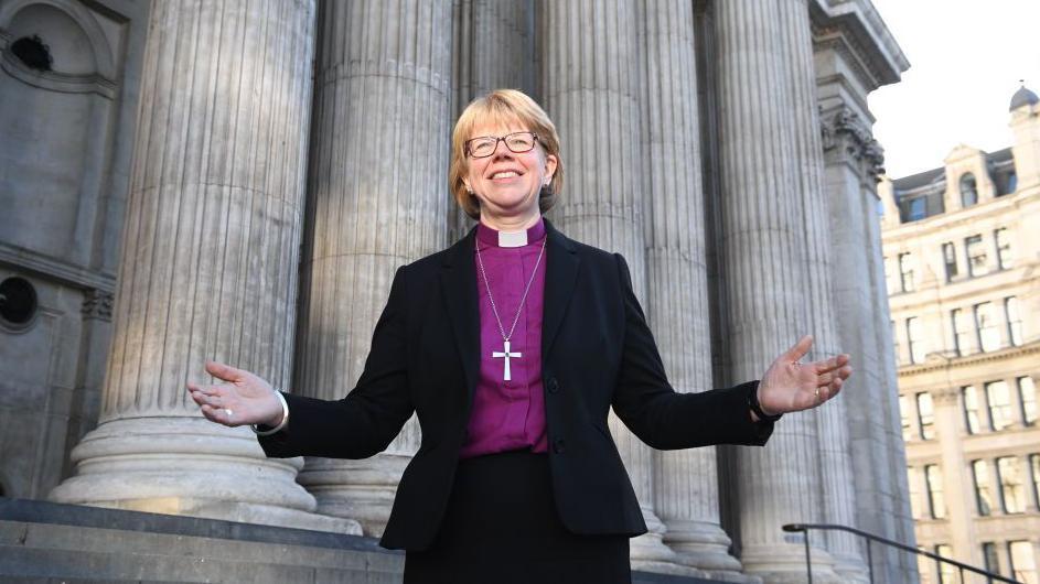 Sarah Mullally con camisa violeta, sonrie abrindo los brazos en frente de la escalinata de la catedral de St. Paul en Londres