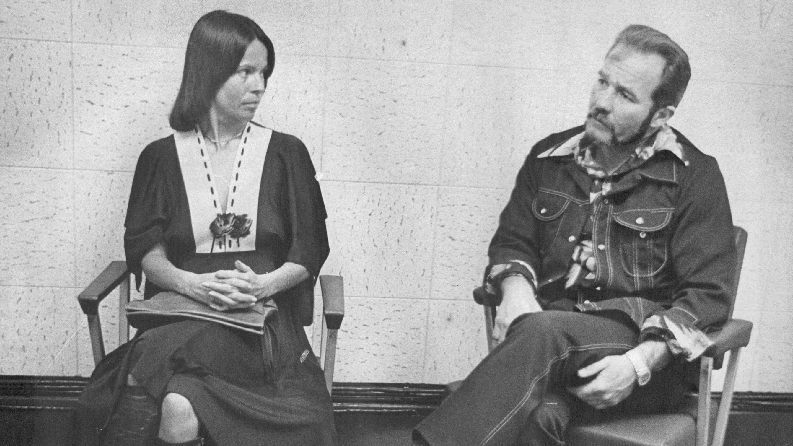 基蒂·奧尼爾（左）坐在椅子上，雙臂交叉，望向達菲·漢布爾頓（右）。她身穿一件深色連衣裙，前襟為淺色V字領。他則穿著深色長領襯衫和牛仔褲。（8/4/1977）