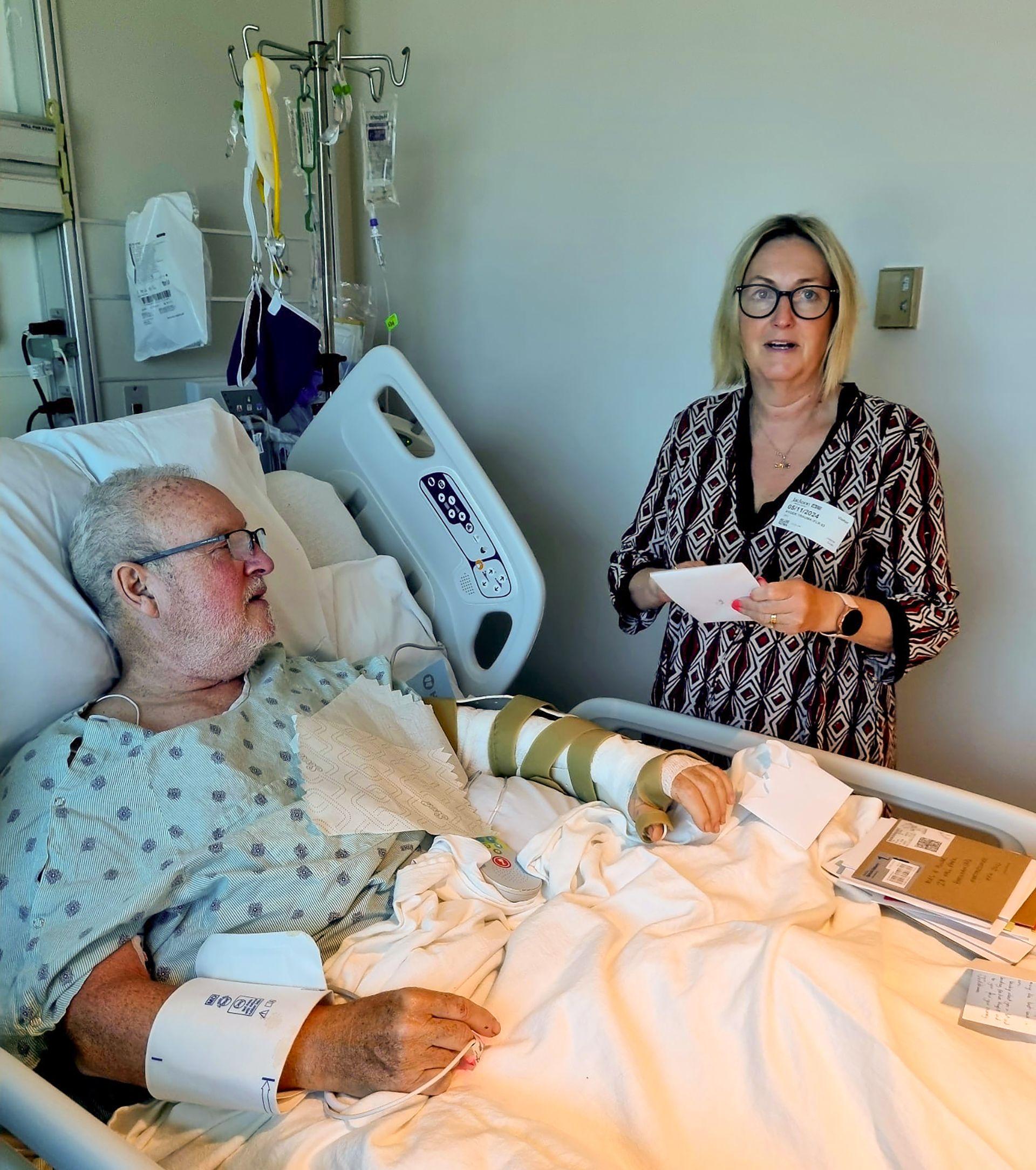 Peter Smith com o bra&ccedil;o esquerdo enfaixado, deitado em uma cama de hospital, com a mulher, Joanna, ao seu lado