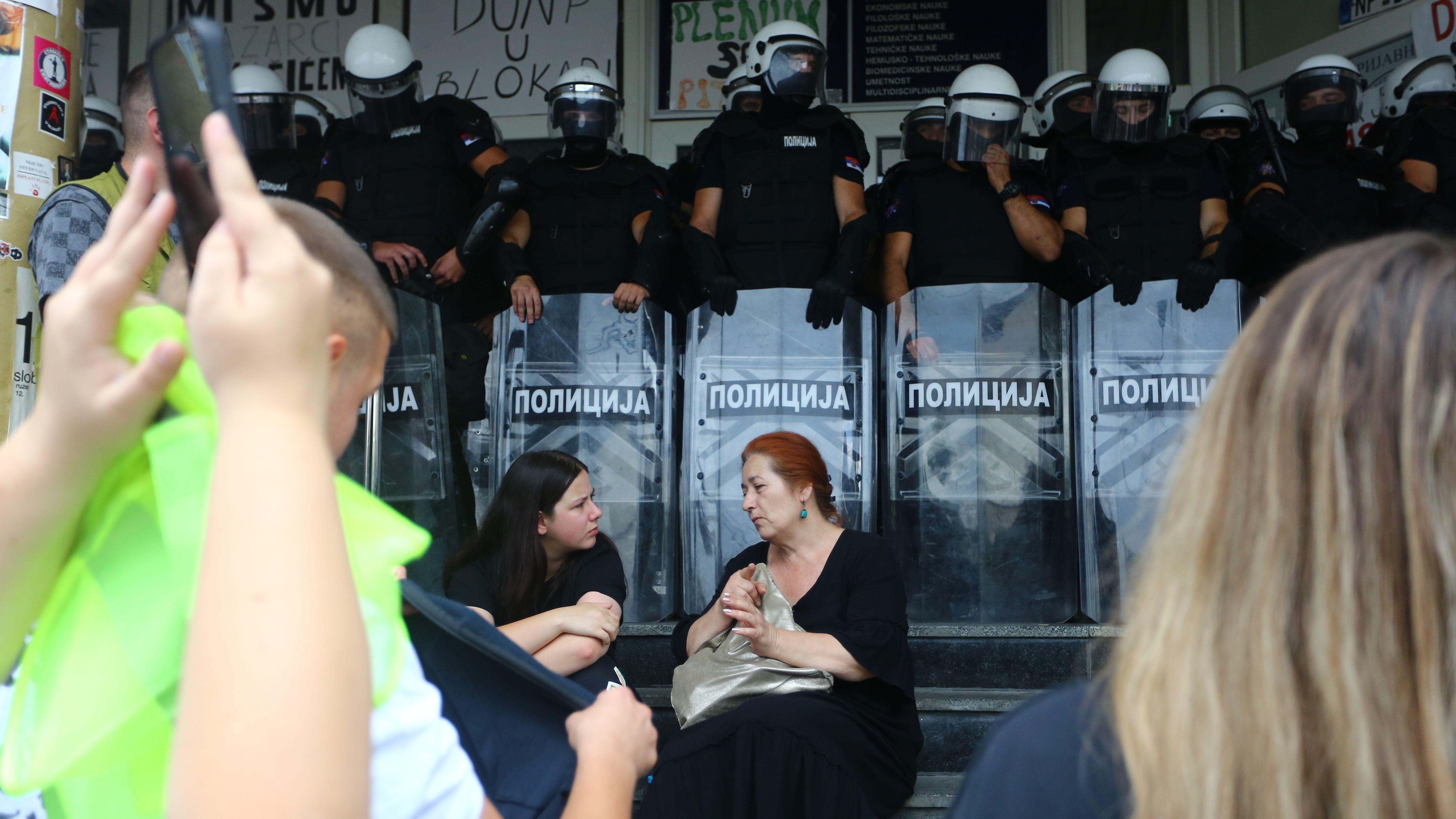 Dve žene, obučene u tamnu odeću sede na stepenicama ispred kordona policije pod punom opremom za razbijanje demonstracija