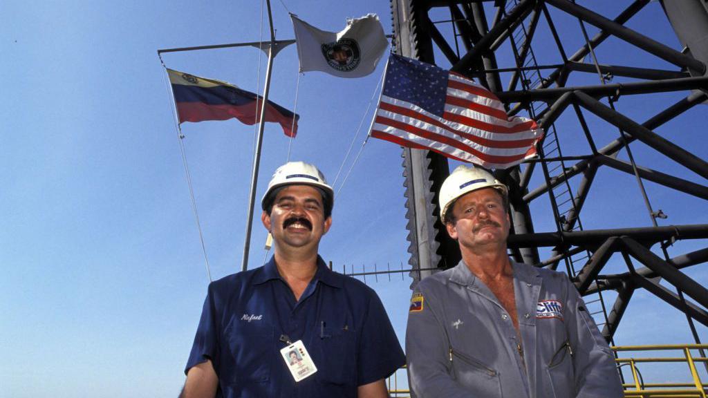 عاملان أمريكي وفنزويلي يقفان أمام منصة لاستخراج النفط في فنزويلا ويبدو خلفها علما كل من الولايات المتحدة وفنزويلا