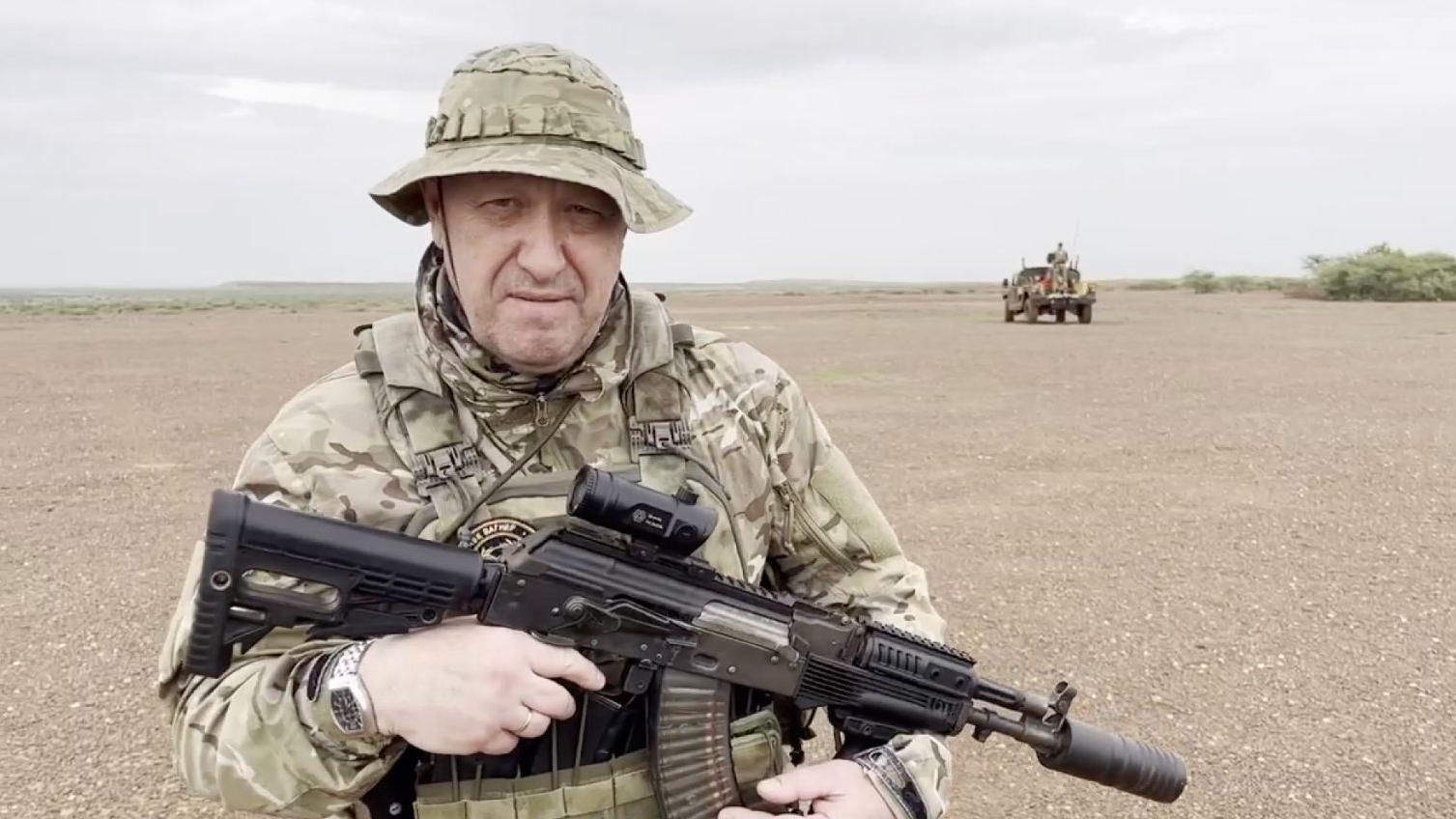 Yevgeny Prigozhin aparece no deserto, vestido com uniforme militar, de chap&eacute;u, segurando uma arma