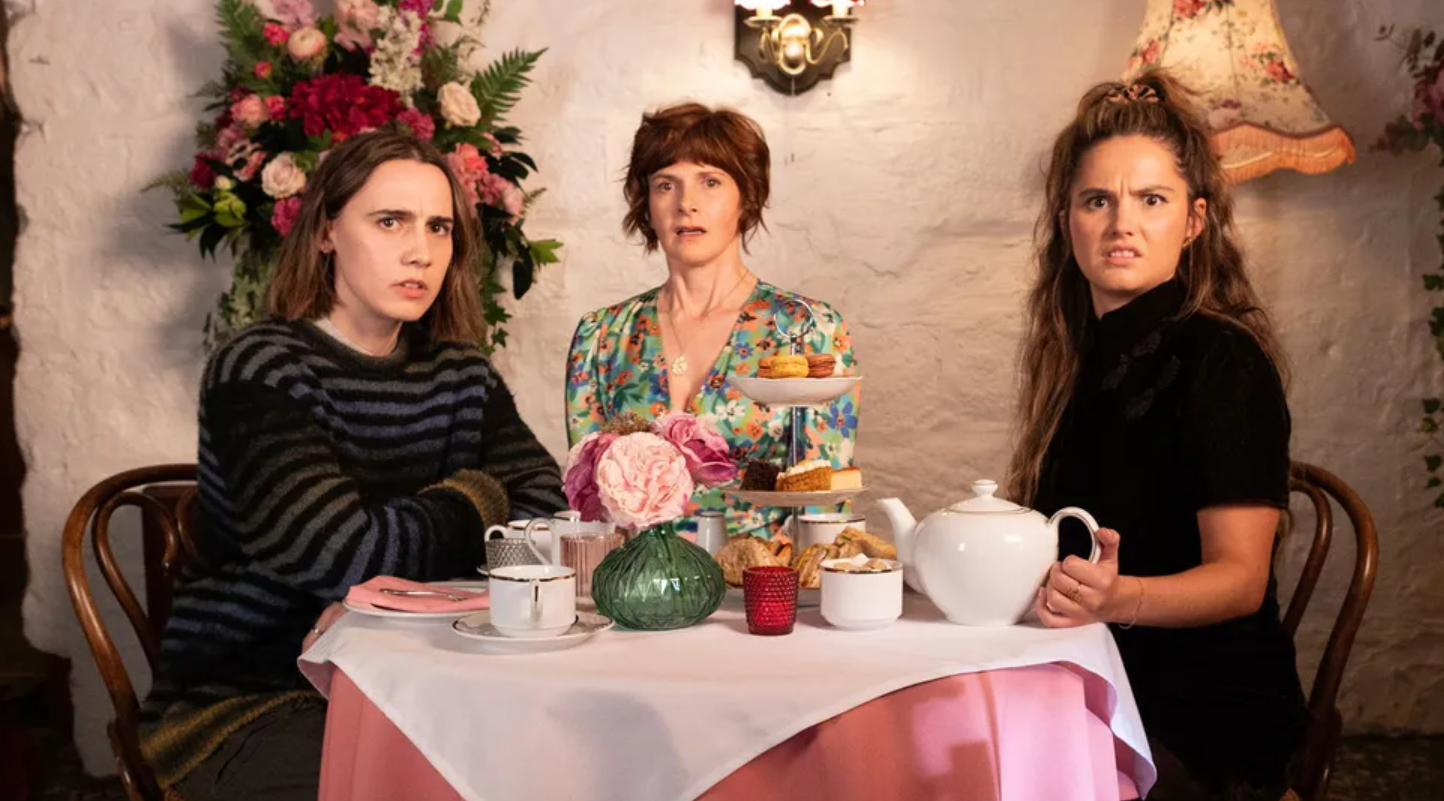 Tres mujeres sentadas a una mesa con tazas y galletas. 