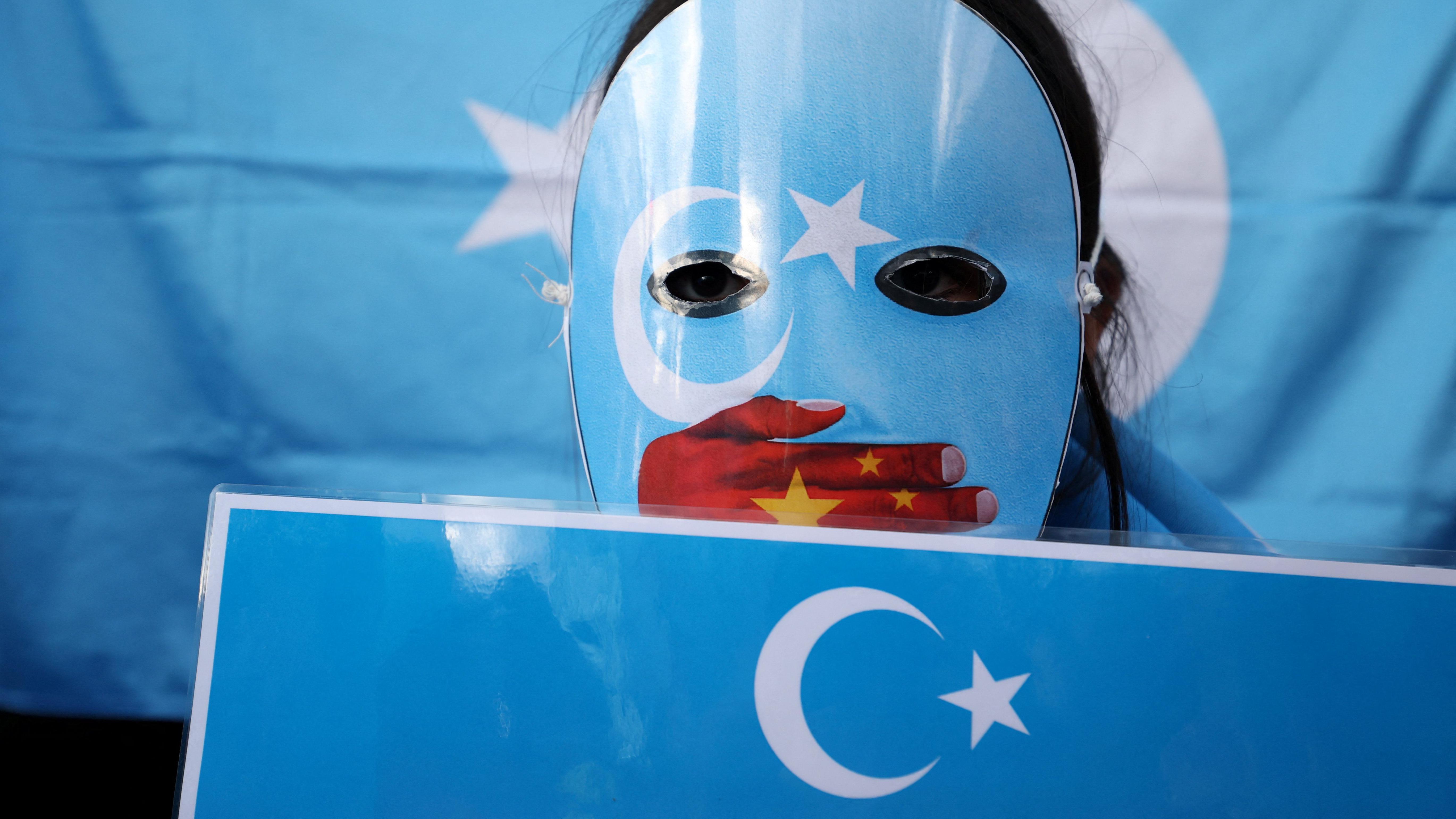 英國倫敦前皇家鑄幣廠中國大使館新館用地前，一位示威者以藍色東突厥斯坦旗面具遮蓋臉龐，面具上畫有一隻手捂住嘴巴圖案，這隻手披上了中國國旗（17/1/2026）