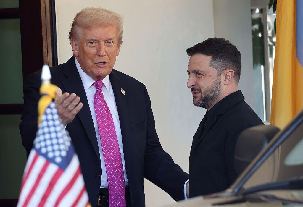 Trump saluda a Zelensky en la entrada de la Casa Blanca y dice algo que parece ser en tono jocoso. Zelensky sonríe. En primer plano se ve la bandera estadounidense en la parte delantera de un coche.