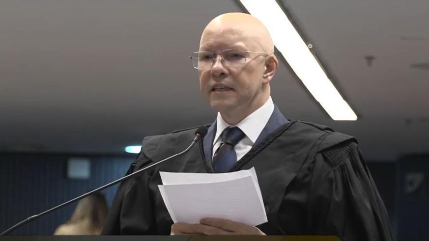 Demóstenes segurando papéis e falando ao microfone em sala de audiência