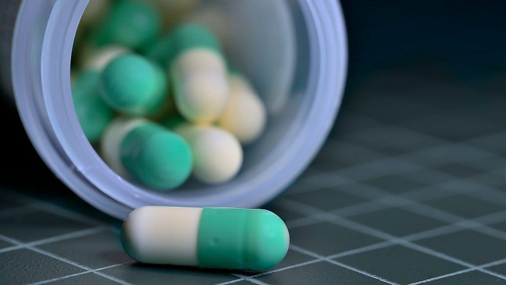 حبوب أدوية بلونين أبيض وأخضر داخل علبة مفتوحة، وهناك حبة واحدة خارج العلبة البلاستيكية الشفافة. على أرضية تميل إل اللون الرمادي ومخطط بالأبيض.