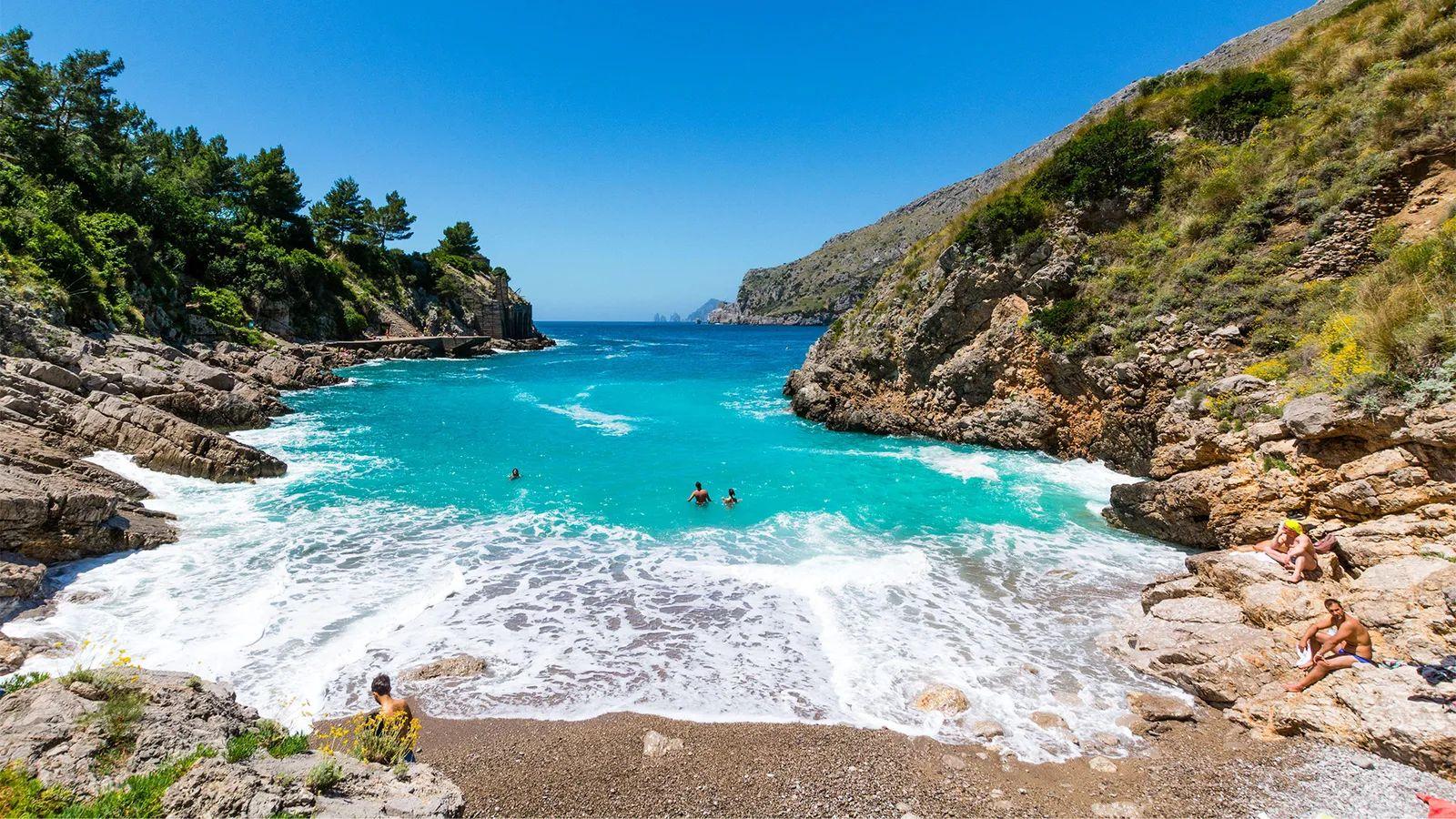 Entrada del mar entre montañas a una pequeña playa de piedras, con tres personas nadando