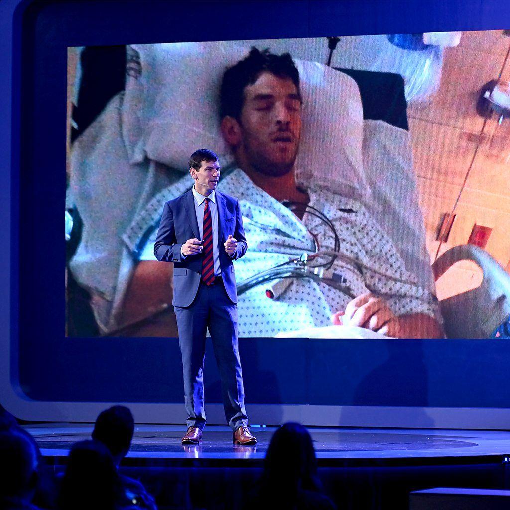 David Fajgenbaum em uma palestra. Ao fundo, imagem de um homem no hospital.