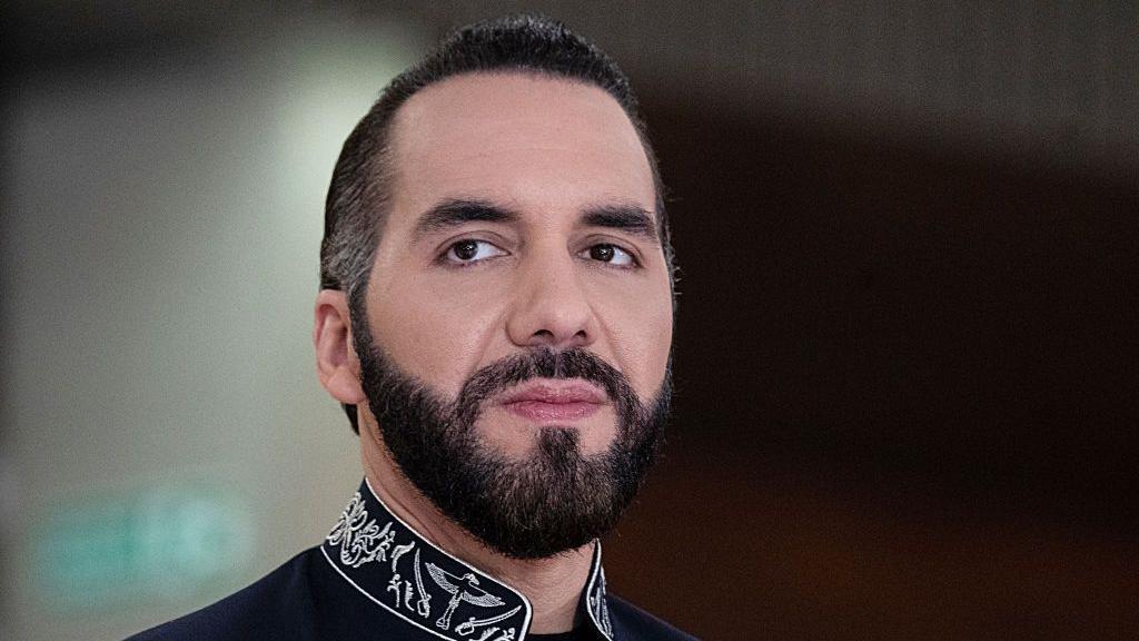 imagen de nayib bukele, presidente de El Salvador. Mira a la cámara con rostro serio. Tiene barba y lleva el cabello hacia atrás. Tiene una camisa negra. 