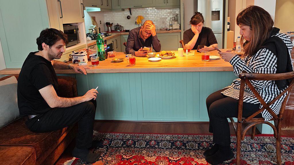 Dos hombres y dos mujeres se sientan alrededor de una mesa de cocina en un apartamento de Teherán revisando sus teléfonos.