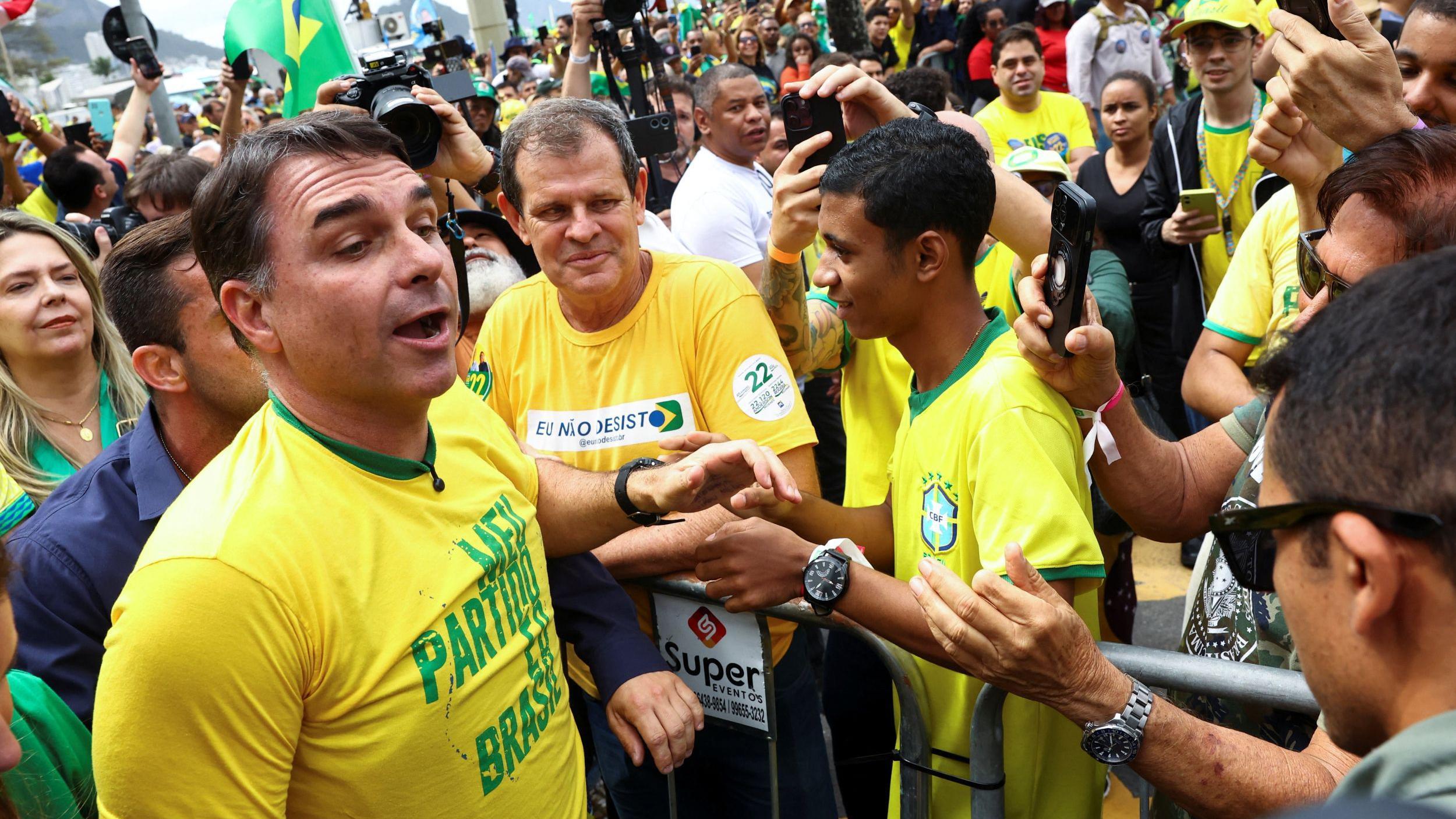 A imagem mostra Fl&aacute;vio Bolsonaro, de camiseta amarela com as cores do Brasil, cercado por apoiadores durante um evento. Ele aparece em primeiro plano diante de uma grade, enquanto pessoas ao redor levantam celulares e c&acirc;meras para registrar o momento; ao fundo, h&aacute; uma grande multid&atilde;o vestida de amarelo e verde, com bandeiras do Brasil.