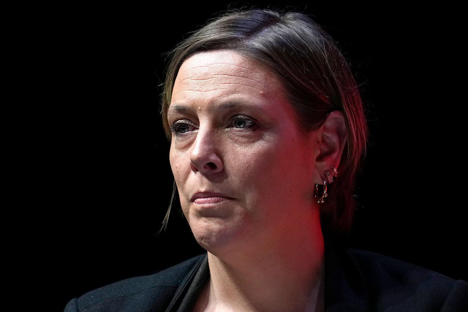 Retrato de Jess Phillips em um palco ou evento com fundo escuro. Ela veste um casaco escuro e usa v&aacute;rios brincos