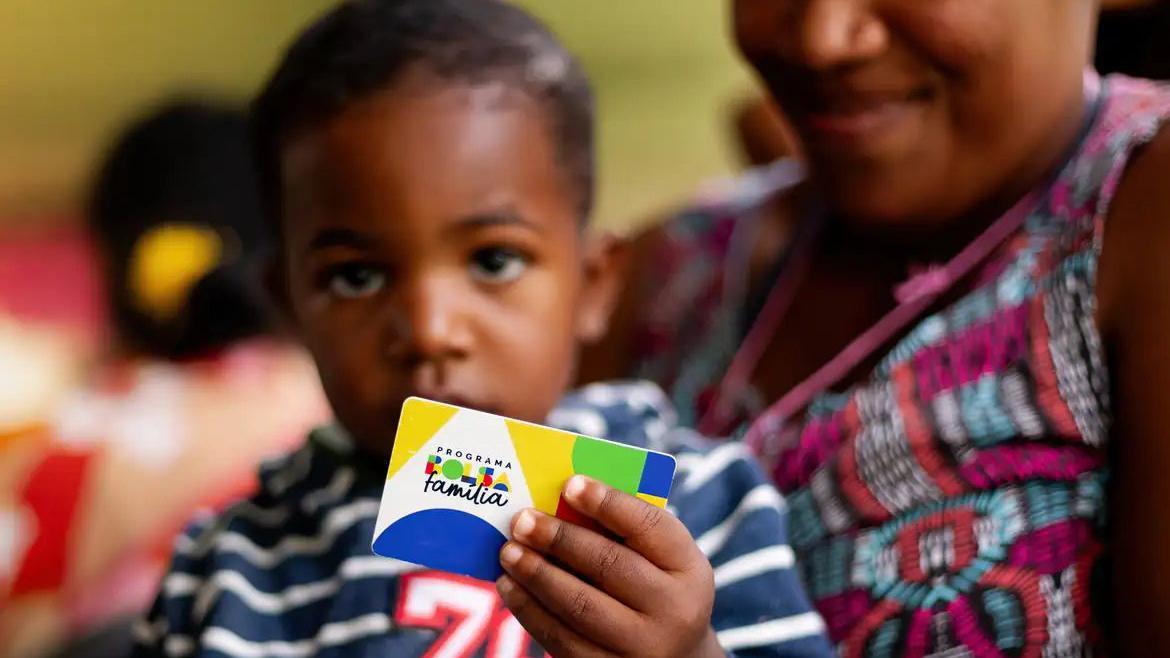 Uma mãe com o filho pequeno no colo e a criança segurando um cartão do Bolsa Família com uma das mãos