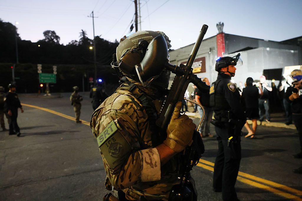Un agente del Servicio de Inmigración y Control de Aduanas (ICE), uniformado, con un arma de gran porte en su mano derecha.
