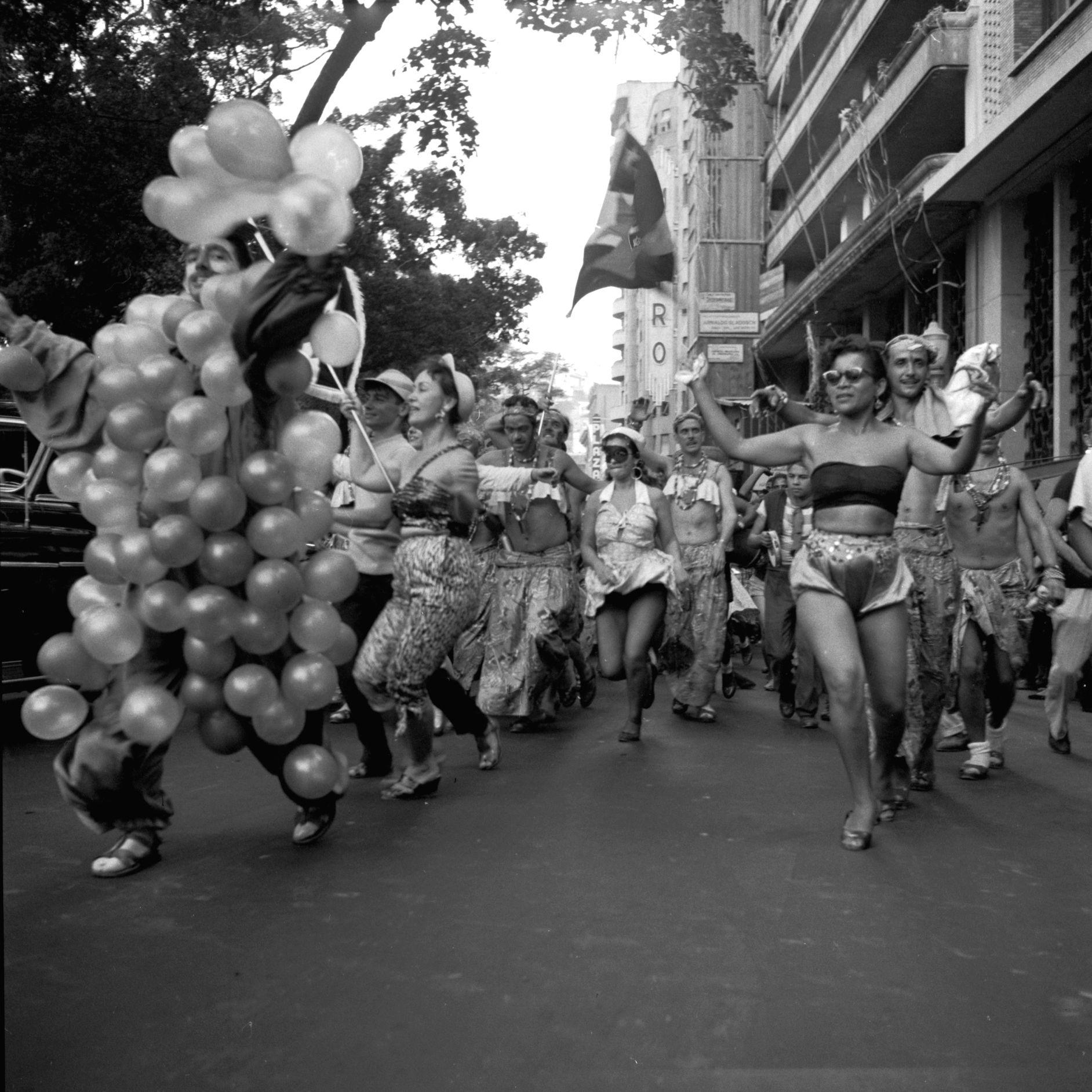 Cena do Carnaval de rua do Rio em 1954: uma pessoa coberta de bal&otilde;es vai &agrave; frente, com outras fantasiadas, carregando estandartes