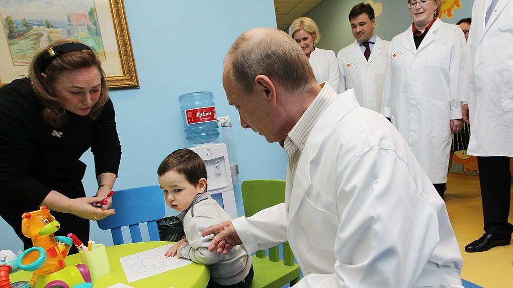 Vladimir Putin, ruski predsednik, u belom mantilu, okrenut ka dečaku koji sedi za malim zelenim stolom