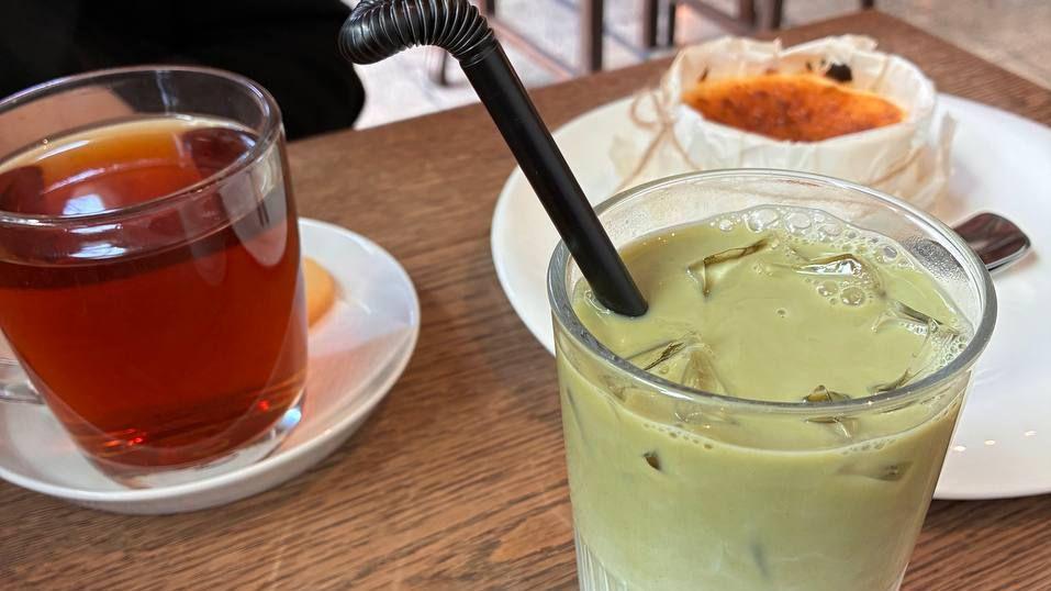 Imagen en primer plano que muestra un matcha latte de color verde en un vaso, junto a una taza de té en otro vaso, que reposa sobre un platillo blanco, con un postre de tarta de queso en un plato al fondo.