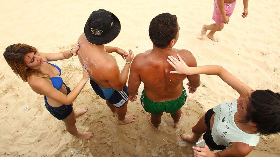 Dos mujeres limpian la espalda de dos hombres con protector solar en una playa. Los hombres llevan shorts de baño, mientras que las mujeres llevan shorts y, en un caso, un top de bikini y en el otro, una camiseta blanca sin mangas.
