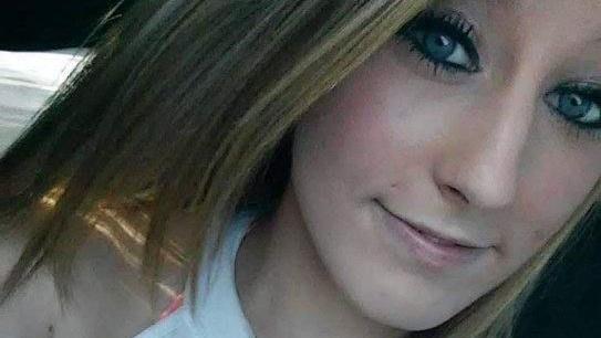 Chelsea Yasser sonríe levemente a la cámara, con su cabello rubio cayéndole sobre la barbilla y los hombros, y sus ojos maquillados con abundante rímel y delineador.