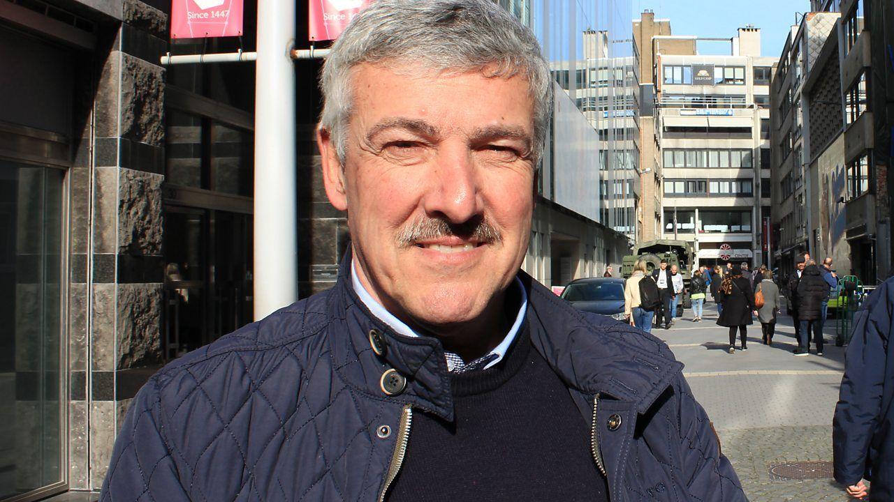 Hombre de bigote y pelo gris con chaqueta acolchada azul de pie en una calle urbana con edificios y transeúntes al fondo
