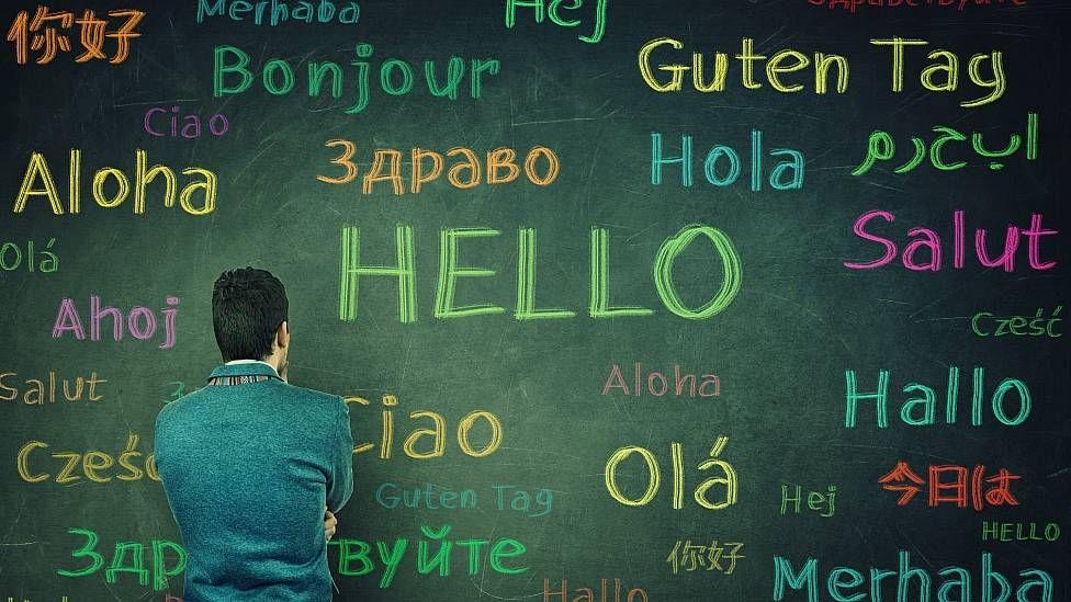 Hombre de espaldas frente a un pizarrón en el que hay escritos saludos en diferentes idiomas