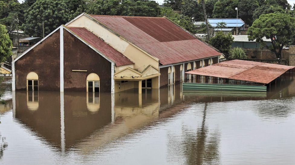 Uma grande casa em Maputo com &aacute;gua da enchente chegando at&eacute; a metade das janelas, com &aacute;rvores atr&aacute;s da casa.