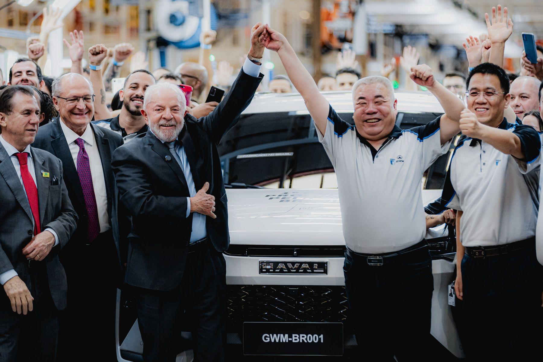 Do lado esquerdo da foto est&atilde;o o presidente Luiz In&aacute;cio Lula da Silva; o vice-presidente e ministro do Desenvolvimento, Ind&uacute;stria, Com&eacute;rcio e Servi&ccedil;os, Geraldo Alckmin; e o ministro do Trabalho e Emprego, Luiz Marinho. Lula segura a m&atilde;o erguida de Mu Feng, CEO da GWM global, que est&aacute; ao lado de Parker Shi, presidente da GWM International, ambos do lado direito da foto. Ao fundo se v&ecirc; um autom&oacute;vel branco, que &eacute; o primeiro ve&iacute;culo produzido pela GWM na nova f&aacute;brica