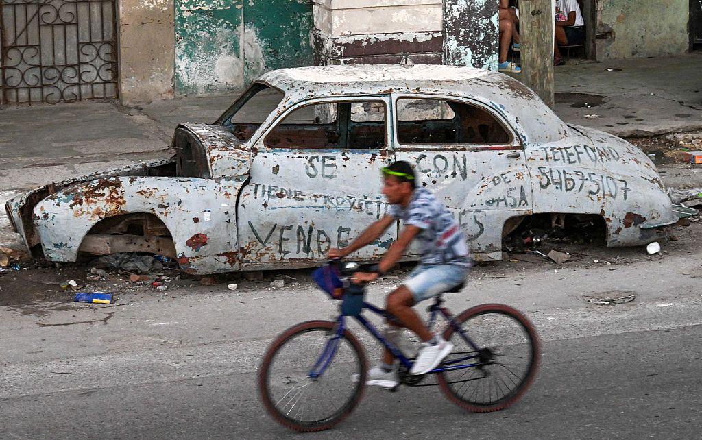 Un hombre maneja una bicicleta junto a un auto destartalado en una calle en Cuba.