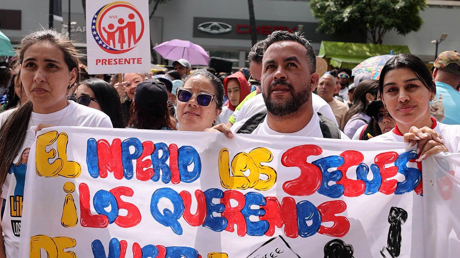 Quatro pessoas carregam uma faixa de protesto. Todos est&atilde;o vestindo camisetas brancas.