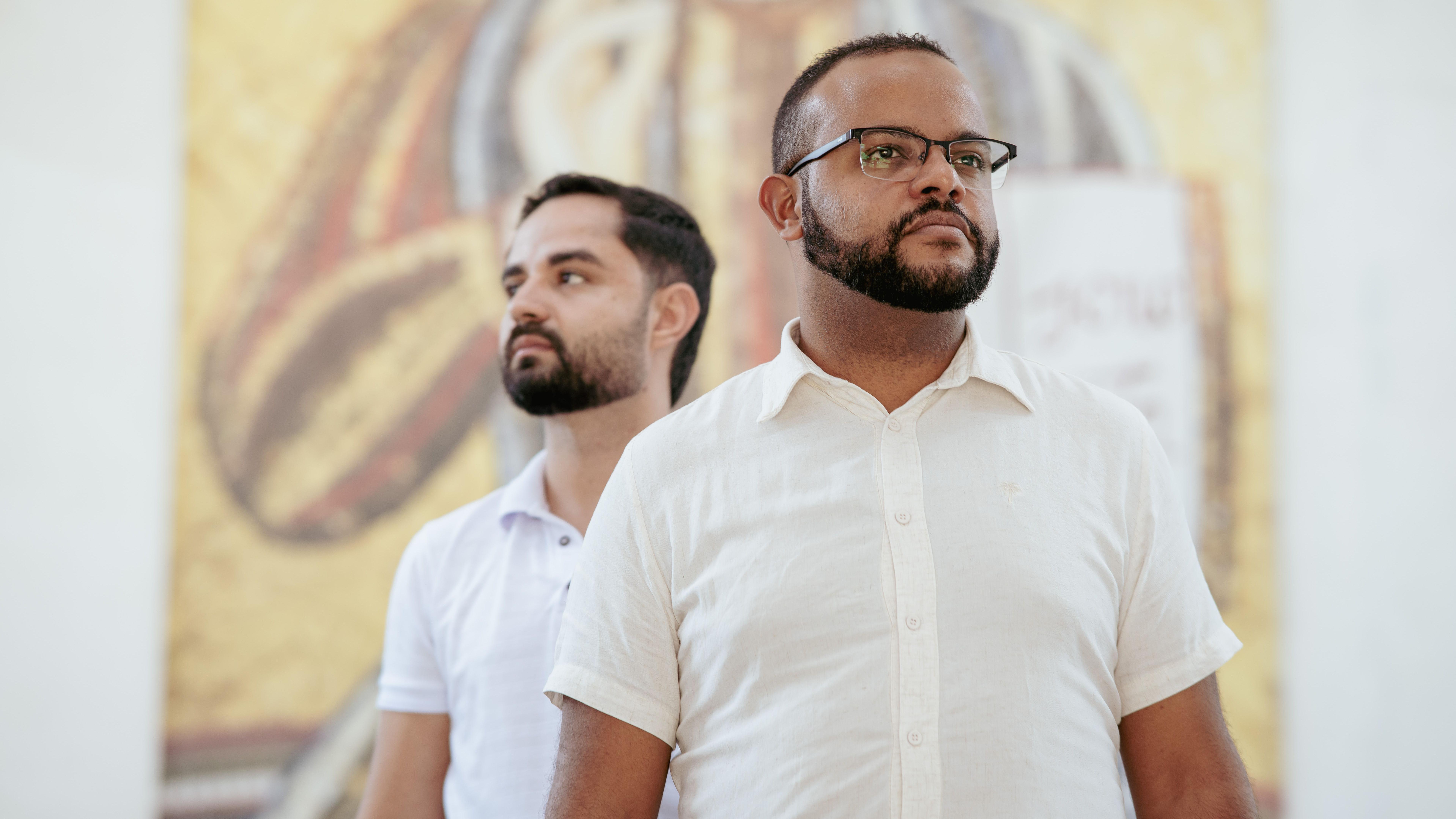Junior Henrique da Silva e Kaik Ribas, ambos de camisa branca de mangas curtas, com um mural religioso ao fundo e olhando ao longe