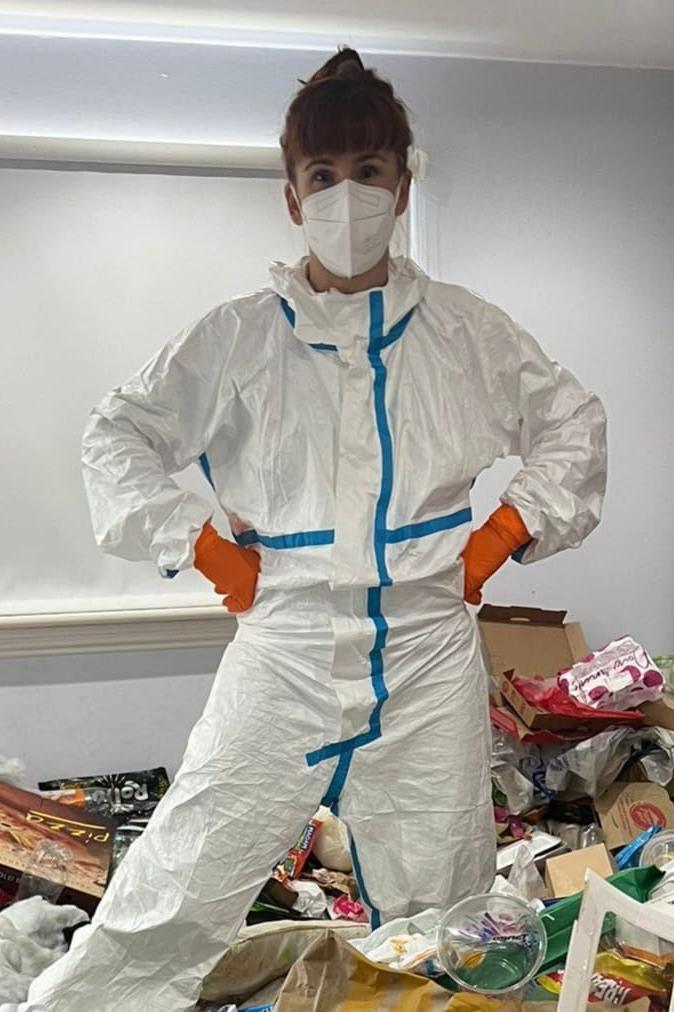 Una mujer de pie sobre un montón de basura dentro de una casa. Tiene el pelo pelirrojo recogido y flequillo. Lleva un traje de protección blanco, una mascarilla blanca y guantes naranjas.