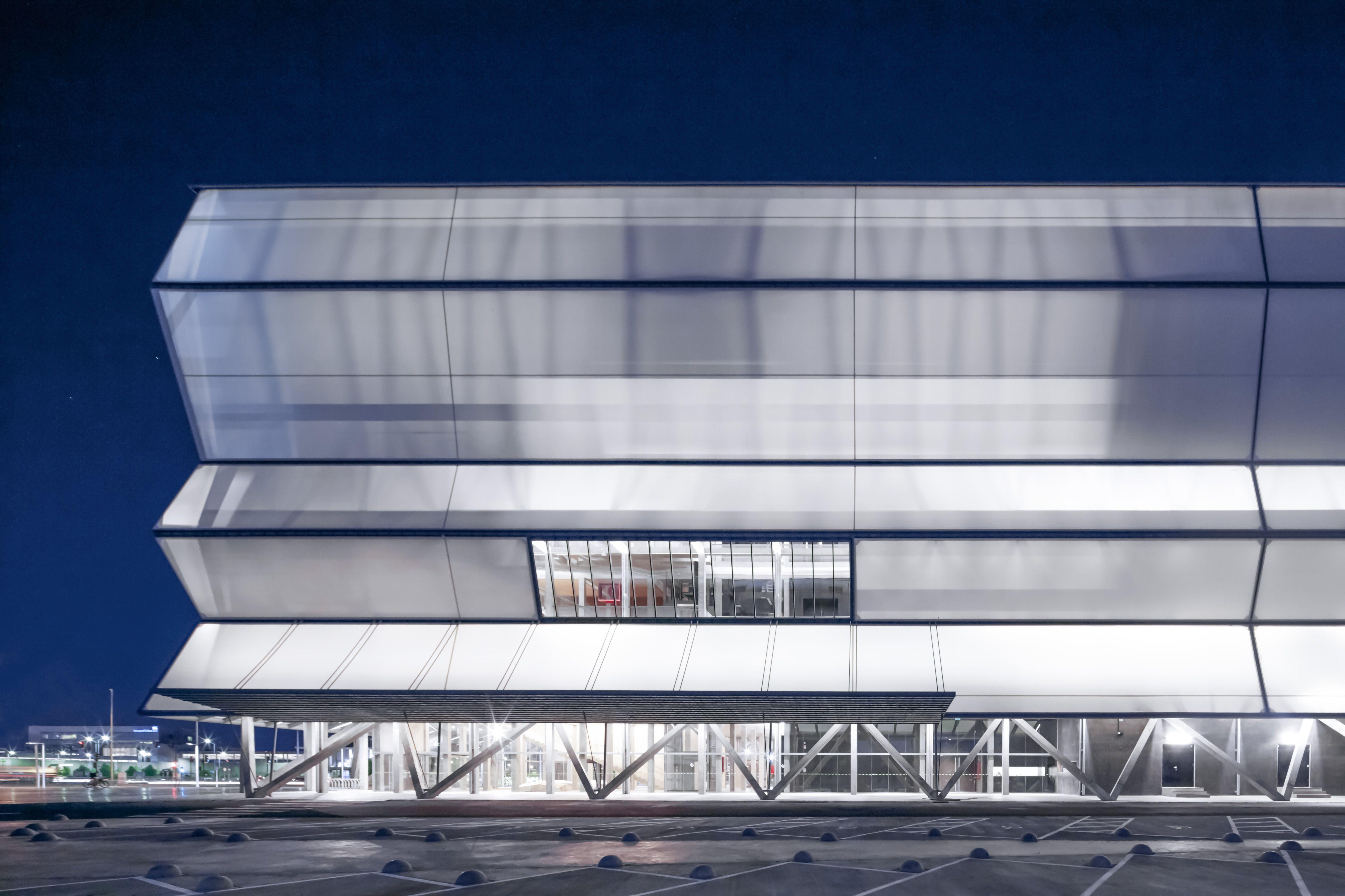 Fachada iluminada de un edificio moderno con paneles superpuestos y estructura metálica visible durante la noche.