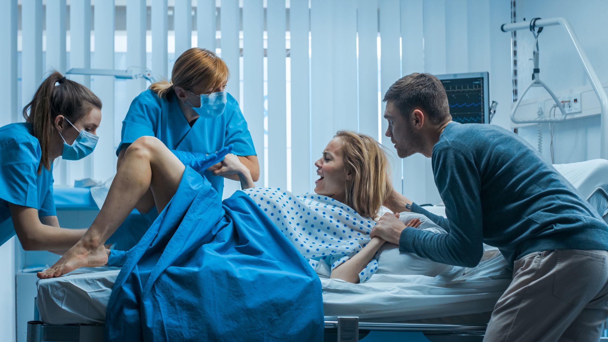 Una mujer dando a luz en un hospital acompañada de su pareja que le toma la mano, mientras dos parteras la asisten